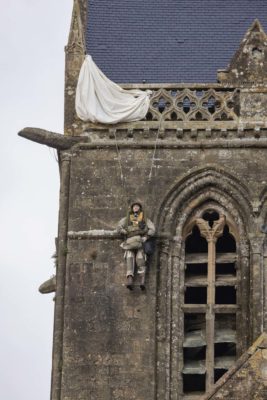 Kirchturm, an dem eine Schaufensterpuppe, kostümiert als Fallschirmspringer, hängt