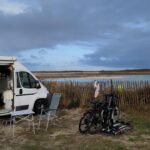 Weißes Womo, Campingmöbel und Fahrräder, vor Staketenzaun, dahinter gleich das Meer