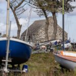 Blick durch ein paar Segelboote an Land, hinter Pinien ein altes Steinhaus